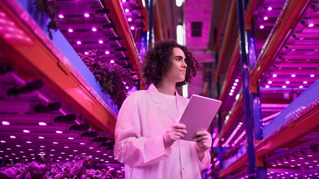 Person in white lab coat and gloves, holding a tablet, standing between rows of vertical farming structures.