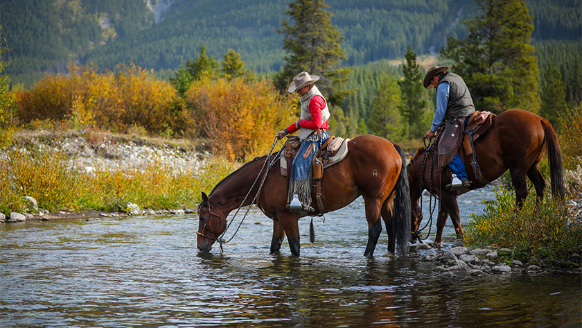 A woman on horseback crossing a river in the foothills of the Rocky Mountains. A man on horseback follows.